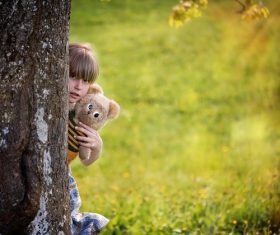 Little girl hugging teddy bear hiding behind tree Stock Photo